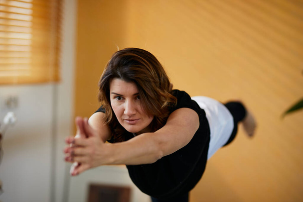 Mujer yogui caucásica de mediana edad de pie en pose de yoga guerrero en casa
. - Foto, imagen