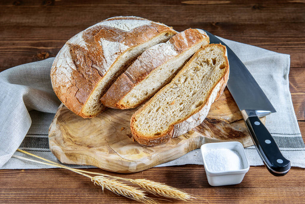 Sliced integral bread, wheat grains, salt and knife on the napkin, on the wooden kitchen table. Healthy homemade integral bread.  - Фото, изображение