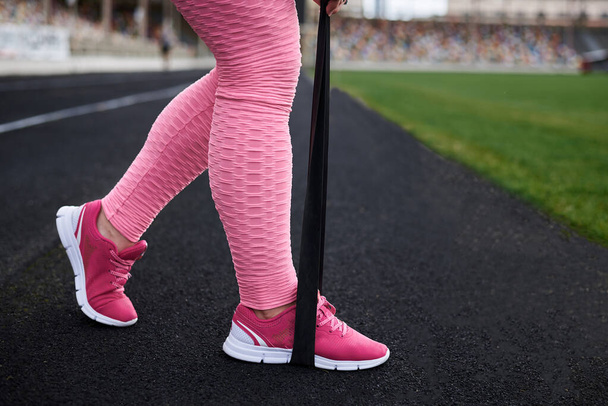 Mujer morena joven, con leggins rosas, entrenando en el estadio en la mañana de verano. Imagen de primer plano de las piernas, haciendo ejercicios de potencia con banda de goma de fitness de resistencia. concepto de estilo de vida saludable
. - Foto, imagen