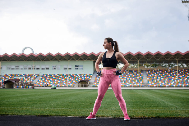 Young brunette woman, wearing pink leggings, sneakers and black top, training on city stadium on summer morning. Sportswoman, stretching, warming up before fitness exercise outside. Healthy lifestyle - Fotografie, Obrázek
