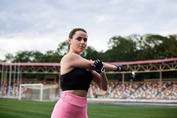 Young brunette woman, wearing pink leggings, sneakers and black top, training on city stadium on summer morning. Sportswoman, stretching, warming up before fitness exercise outside. Healthy lifestyle - Fotó, kép