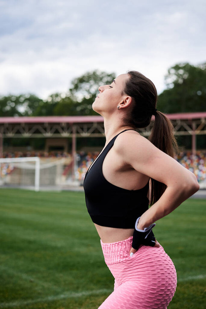 Young brunette woman, wearing pink leggings, sneakers, black top and fitness gloves, training on city stadium in summer. Sportswoman, stretching, warming up before exercise outside. Healthy lifestyle - Foto, Bild