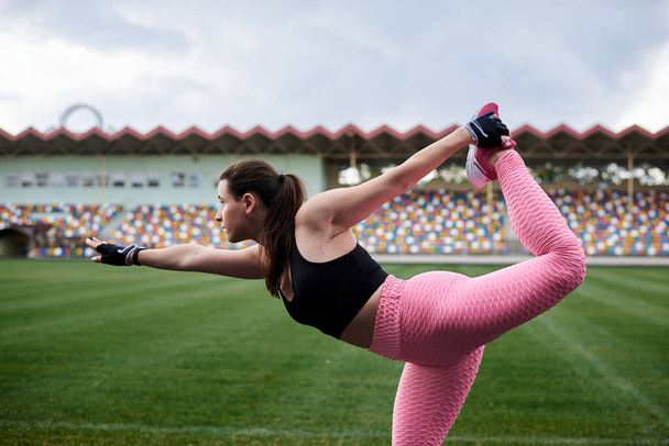 Young brunette woman, wearing pink leggings, sneakers, black top and fitness gloves, training on city stadium in morning. Sportswoman, stretching, warming up before exercise outside. Healthy lifestyle - Foto, Imagen