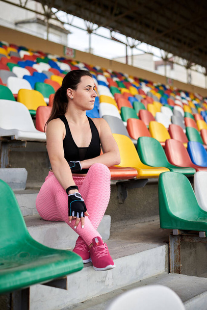 Young brunette sportswoman, wearing black top, pink leggings and sneakers, sitting on colorful seats on stadium tribune, relaxing after training, thinking. Active healthy lifestyle concept. - Фото, зображення