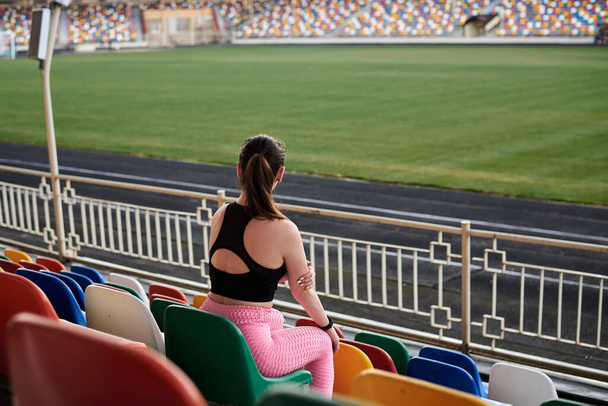 Young brunette sportswoman, wearing black top, pink leggings and sneakers, sitting on colorful seats on stadium tribune, relaxing after training, thinking. Active healthy lifestyle concept. - Photo, Image