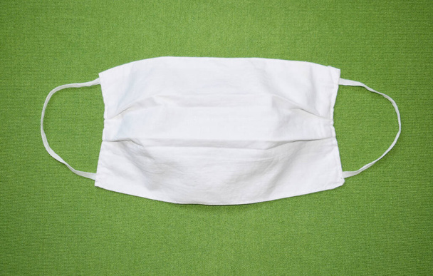 Wit beschermend gezichtsmasker gemaakt van stof op een groene achtergrond. Masker met elastische banden aan de zijkanten. Uitzicht van bovenaf. - Foto, afbeelding