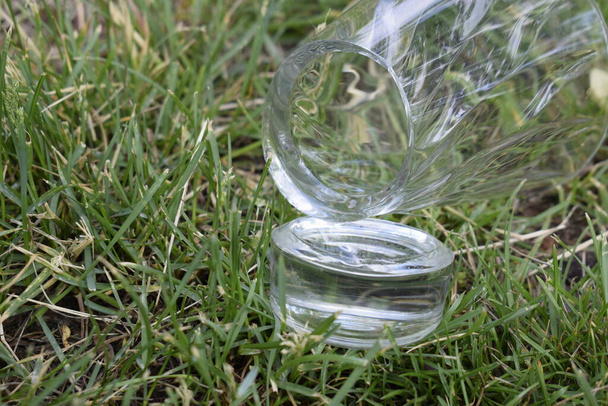 Разбитая стеклянная чашка на зеленой траве в саду. Разбитое толстое дно стекла
. - Фото, изображение
