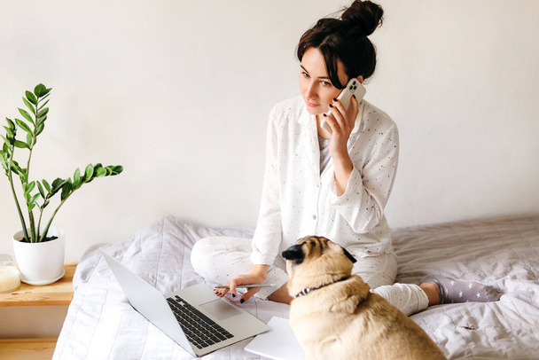 Young beatiful girl is talking by phone and writes a note and near her is lying a dog. Home office. Concept of working and studying at isolation. - Φωτογραφία, εικόνα
