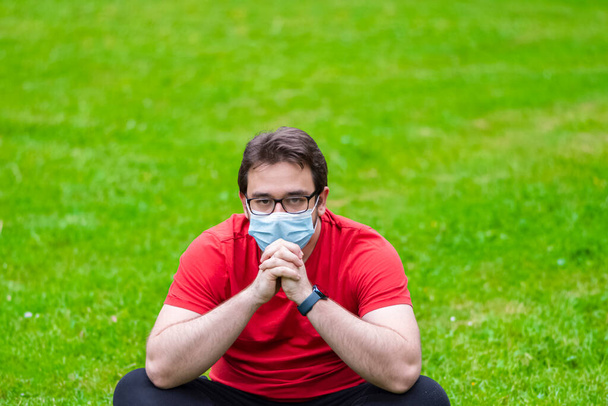 練習後に休息を取るために草の上に座ってコロナに対する措置に従うためにフェイスマスクを身に着けている脂肪男 - 写真・画像