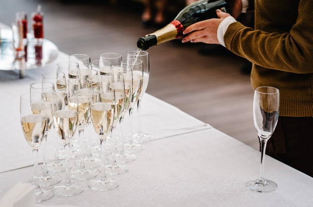 Man hands pouring champagne from a bottle in glass that stand on white table. Close up. The man fills the glasses elite drinks standing in three rows. - Fotografie, Obrázek