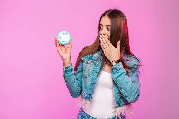 Trendy young woman in stylish denim blue jacket in white T-shirt closes mouth with hand and looks at funny donut in a medic mask. Girl posing near pink wall indoors. Funny concept virus covid-19. - Foto, afbeelding