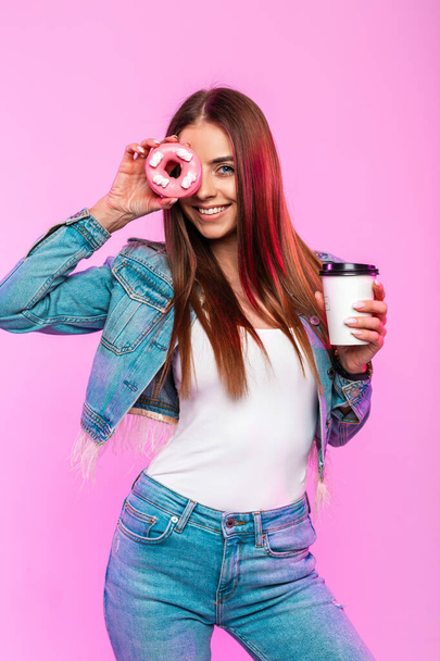 Радостная молодая женщина с положительной улыбкой и чашкой чая держит вкусный пончик в глазури возле лица в помещении. Веселая девушка в моде синяя джинсовая одежда наслаждается десерт возле розовой стены в студии
. - Фото, изображение