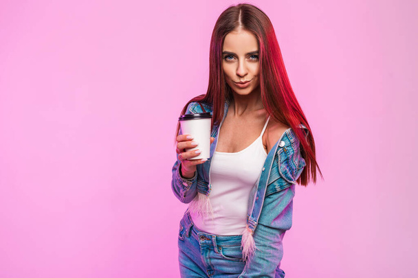 Joyful attractive young woman with positive smile in fashionable youth jeans clothes with cup of tea poses and cute smiles near modern pink wall in the studio. Happy girl enjoys coffee break indoors. - Photo, Image