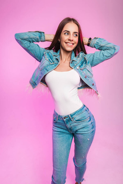 Fröhliches Mode-Model junge Frau in Jeansjacke in Vintage-Jeans in stilvollem T-Shirt posiert lächelnd neben moderner pinkfarbener Wand drinnen. Hübsches positives Mädchen mit süßem Lächeln in trendigen Klamotten im Zimmer. - Foto, Bild