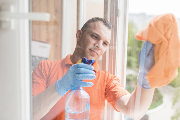 Ο τύπος με τα γάντια και τις στολές από την καθαριότητα πλένει το παράθυρο με ένα πανί και σπρέι. - Φωτογραφία, εικόνα