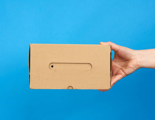 hand houdt een bruine kartonnen doos van papier op een blauwe achtergrond, close-up - Foto, afbeelding