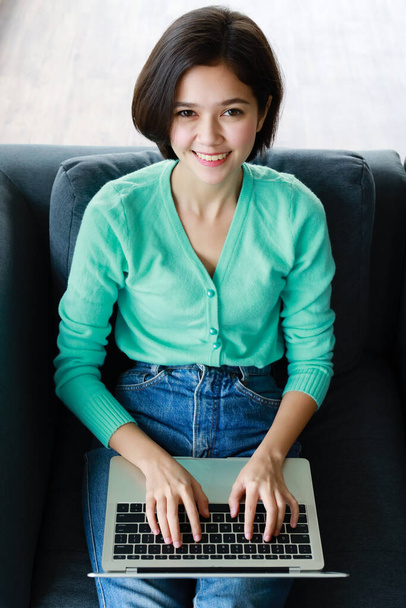 Молодая, симпатичная и красивая пол-расы тайско-немецкая девушка сидит и улыбается, как стол, в то время как использует настольный компьютер, работающий на диване в гостиной. Идея для счастья молодые люди учатся и учатся на дому. - Фото, изображение