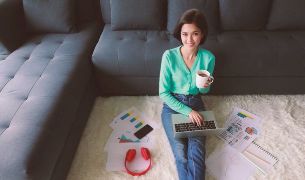 Young and beautiful Asian woman holding coffee cup and sitting in house beside sofa with easy and cozy manner using laptop notebook computer to work and study in private area. - Foto, imagen