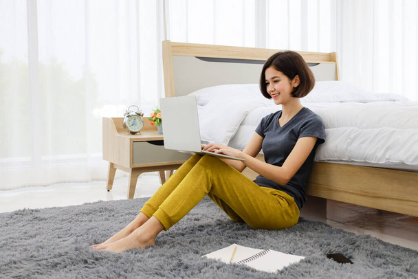Beautiful young teen half Thai-German girl sitting beside bed in bedroom using laptop notebook computer working with coffee cup in cozy house room. Idea for work from home lifestyle of modern woman. - Photo, Image
