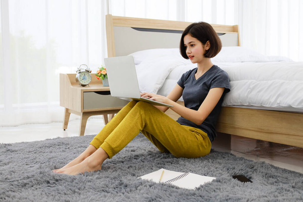 Krásná mladá dospívající polovina thajsko-německé dívka sedí vedle postele v ložnici pomocí notebooku počítače pracuje s šálkem kávy v útulném pokoji. Nápad na práci z domova životní styl moderní ženy. - Fotografie, Obrázek