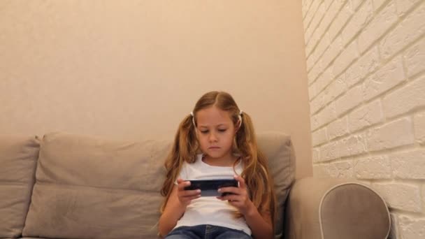 Zwykłe dziecko siedzące na kanapie w domu i dotykające telefonu komórkowego - Materiał filmowy, wideo