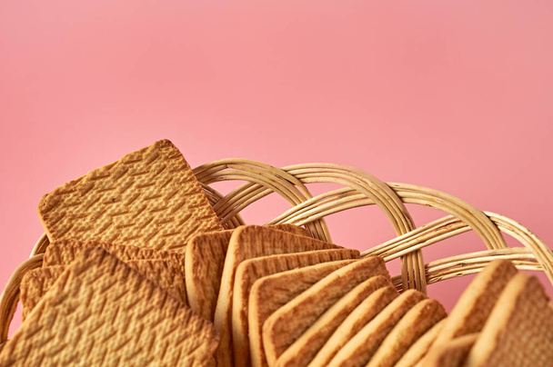Wooden wicker basket full of whole square cookies lies on pink desk on kitchen. Space for text - Foto, Bild