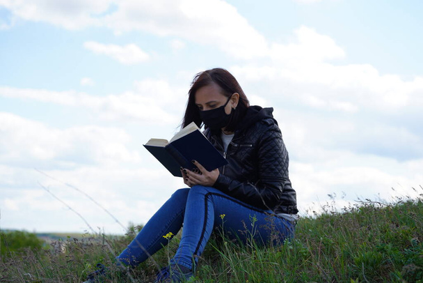 Lähikuva nuori nainen musta lääketieteellinen naamio ja rento vaatteita istuu mäellä ja lukeminen kirja pilvinen päivä. Aikuinen nainen lepää yksin maaseudulla - Valokuva, kuva