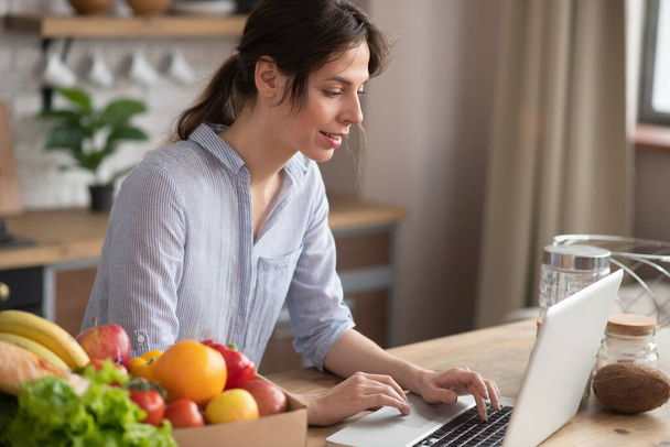 Girl in grey homewear sitting in the kitchen and reading something online - Zdjęcie, obraz