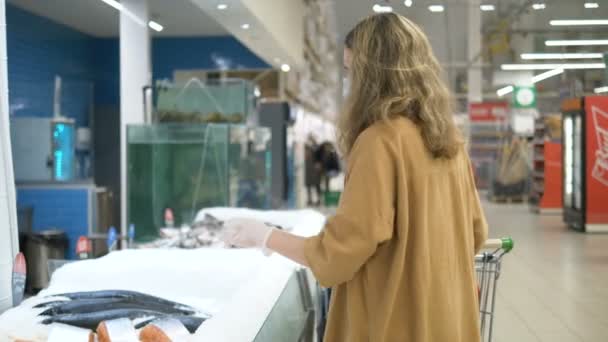 医療マスクと手袋のスーパーマーケットの若い女の子は魚を購入します。コロナウイルス感染に対する注意事項. - 映像、動画