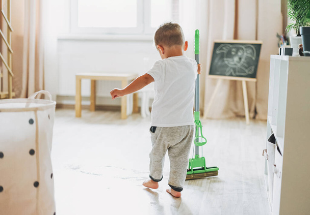 Cute baby boy with mop cleaning floor at room. Child wipes water in bright interior - Foto, immagini
