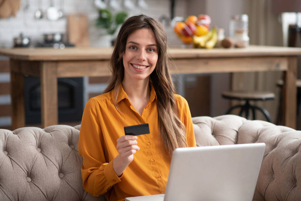 Long-haired girl in a mustard color blouse holding a credit card and smiling - Фото, зображення