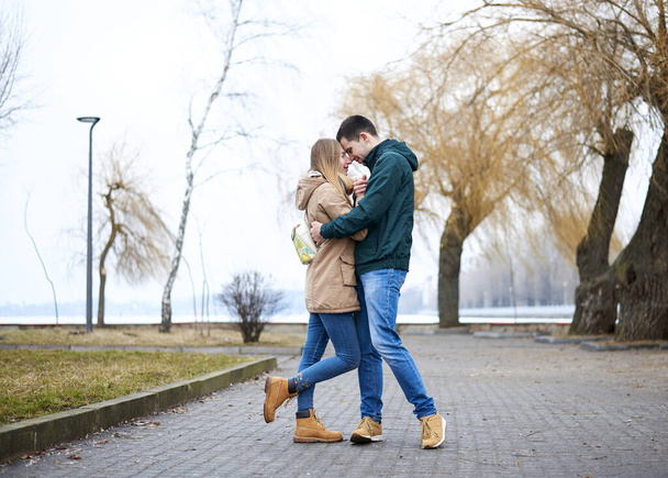 Young couple in love, standing, embracing hugging each other, wearing casual clothes and jeans, on the rainy spring day in city park. Romantic Valentines day celebration outside in town. - Photo, Image
