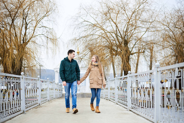 Young couple in love, wearing beige and green casual jackets, holding hands, walking on the lock bridge in winter with yellow trees on the background. Romantic Valentines day celebration. - Photo, Image