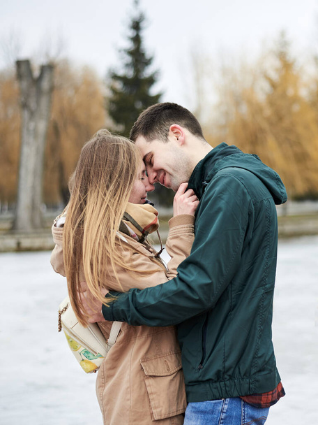 Young couple in love, wearing beige and green casual jackets, standing in front of frozen lake hugging, kissing each other in winter. Valentines day romantic celebration outside - Fotoğraf, Görsel