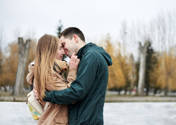 Young couple in love, wearing beige and green casual jackets, standing in front of frozen lake hugging, kissing each other in winter. Valentines day romantic celebration outside - Foto, Bild