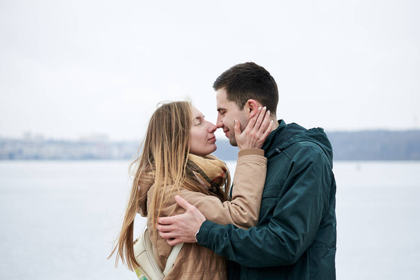 Mladý zamilovaný pár, v béžových a zelených neformálních bundách, stojí před zamrzlým jezerem, objímají se a líbají se v zimě. Valentýn romantické oslavy venku - Fotografie, Obrázek