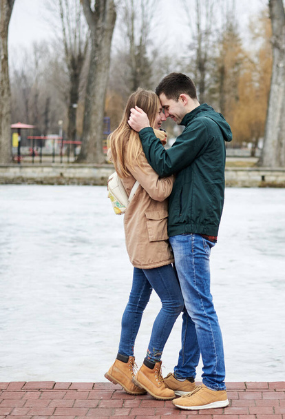 Young couple in love, wearing beige and green casual jackets, standing in front of frozen lake hugging, kissing each other in winter. Valentines day romantic celebration outside - Fotó, kép