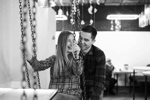 Young couple in love, wearing red checkered shirts, sitting on a swing in coffee shop. Attractive brunette man swinging pretty blond woman in coffee shop, smiling. Romantic Valentines day celebration - Zdjęcie, obraz