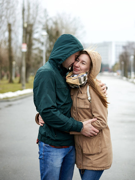 Young couple in love, standing on the park path, wearing hoods, hugging, looking at each other on the rainy spring day. Romantic Valentines day celebration - Photo, image