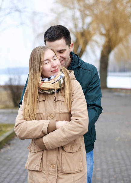 Young brunette man, wearing green jacket, standing behind young blond woman, wearing beige jacket, hugging her, smiling, kissing. Romantic Valentines day celebration - Фото, изображение