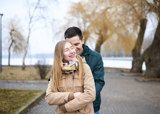 Young brunette man, wearing green jacket, standing behind young blond woman, wearing beige jacket, hugging her, smiling, kissing. Romantic Valentines day celebration - Фото, изображение