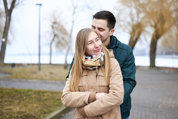 Young brunette man, wearing green jacket, standing behind young blond woman, wearing beige jacket, hugging her, smiling, kissing. Romantic Valentines day celebration - Foto, afbeelding
