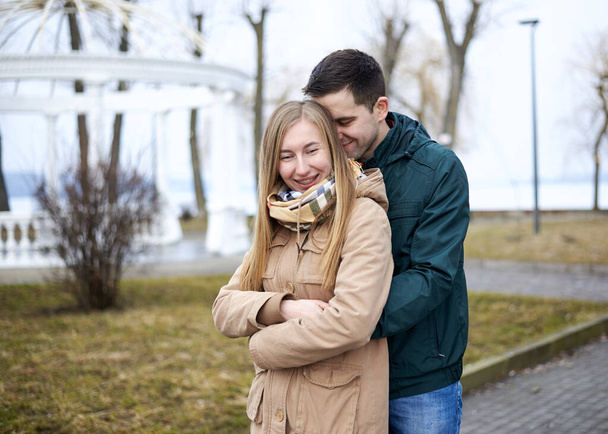 Young brunette man, wearing green jacket, standing behind young blond woman, wearing beige jacket, hugging her, smiling, kissing. Romantic Valentines day celebration - Photo, Image
