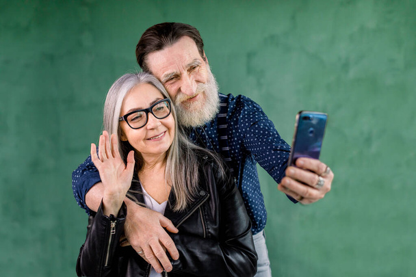Portrait of attractive senior couple, standing at home in front of green wall and talking with their friends via phone video chat. Bearded man embracing his wife while she waving hand to phone camera - Φωτογραφία, εικόνα