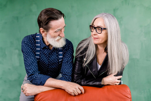 Romantic senior couple, posing in stylish room on green wall background. Cheerful smiling senior couple, bearded man and charming gray haired lady, leaning on red soft armchair and looking each other - Фото, изображение