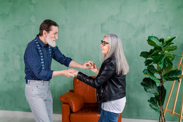 Beautiful joyful stylish senior woman with long straight gray hair, dancing together with her handsome bearded husband in the cozy room in front of green wall - Foto, Bild