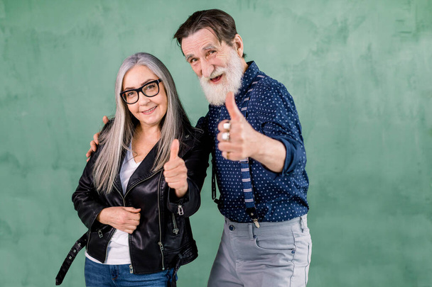 Lovely senior smiling joyful couple, handsome bearded man and pretty gray haired woman, posing to camera in front of green wall and showing their thumbs up gesture. - Photo, Image