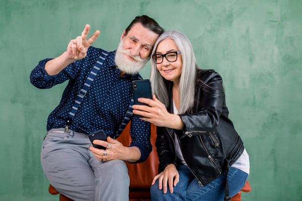 Attraktives stylisches Paar, bärtiger Mann und grauhaarige Dame, die zusammen auf dem roten Stuhl sitzen, die Handykamera zum Fotografieren benutzen und lächeln. Isoliert auf grünem Hintergrund. - Foto, Bild