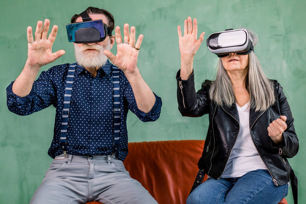 Front view of excited cheerful elderly man and woman, sitting together on red soft chair on green background, and using vr glasses headset, touching imaginary screen in the air - Foto, immagini