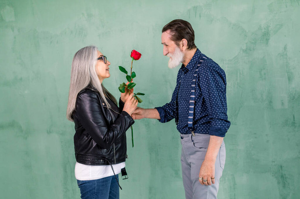 Happy senior family portrait. Handsome elderly bearded man giving beautiful red fresh rose to his charming smiling wife with long straight gray hair, standing on green wall background - Photo, Image